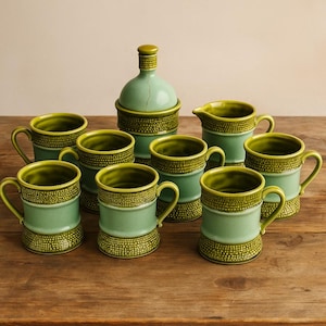 Könnte beinhalten: Ein Set aus grünen Keramikbechern, einem Sahnekännchen und einer Zuckerdose mit Deckel. Die Becher haben einen Henkel und ein strukturiertes Band. Zuckerdose und Sahnekännchen haben ein ähnliches Design. Das Set steht auf einer Holzoberfläche.