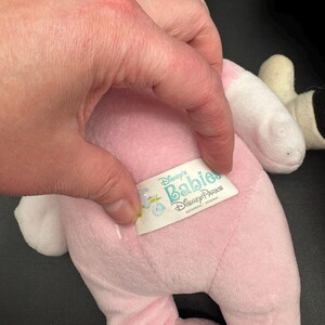 May include: A pink plush toy with white accents, featuring a tag that reads "Disney's Babies Disney Parks". The toy is held by a hand with a silver ring. The toy appears to be a baby version of a Disney character.