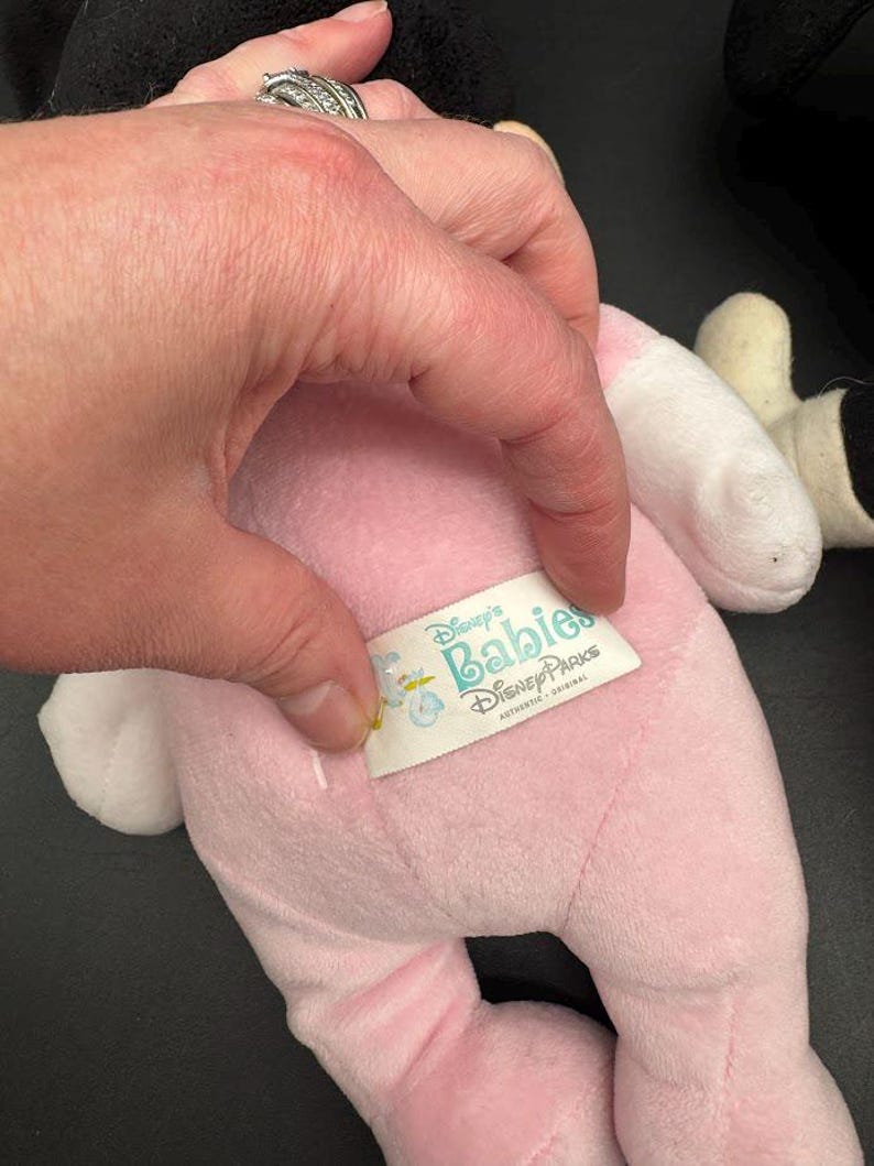 May include: A pink plush toy with white accents, featuring a tag that reads "Disney's Babies Disney Parks". The toy is held by a hand with a silver ring. The toy appears to be a baby version of a Disney character.