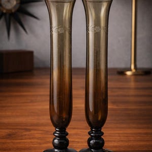 Op de afbeelding: Twee hoge, bruine glazen vazen met een bloemen geëtst ontwerp aan de bovenkant. De vazen hebben een uitlopende bovenkant en een zwarte, afgeronde basis. De vazen staan op een houten oppervlak.