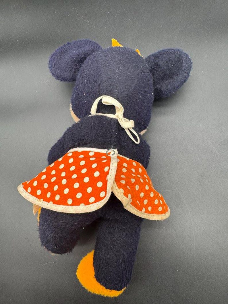 May include: A plush toy mouse wearing a red and white polka dot skirt. The mouse is black with yellow ears and feet.