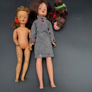 May include: Three vintage dolls are shown against a dark backdrop. One doll is nude, another is wearing a black and white checked dress. A third doll's head and hair are separated from its body.