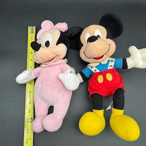 May include: Two plush Disney character dolls. One is Minnie Mouse, wearing a pink outfit and bow. The other is Mickey Mouse, wearing red shorts, yellow shoes, and a blue and white top. A measuring tape is in the image.