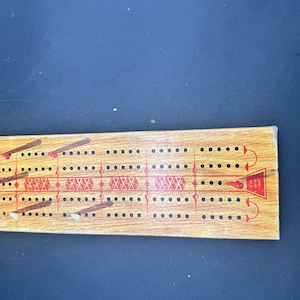 Può includere: Tavola da cribbage in legno con segni rossi e neri. La tavola ha più file di fori per l'inserimento dei pioli e un disegno rosso al centro. I pioli sono marroni e bianchi. Il testo "STANDARD CRIBBAGE BOARD" è visibile.