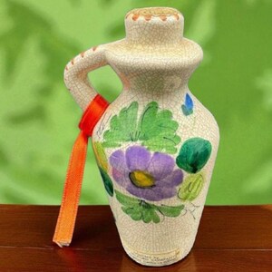 May include: A cream ceramic jug with a handle and spout, decorated with a purple flower, green leaves, and an orange ribbon tied around the handle. The jug has a crackle glaze finish. The jug is likely used for pouring liquids.