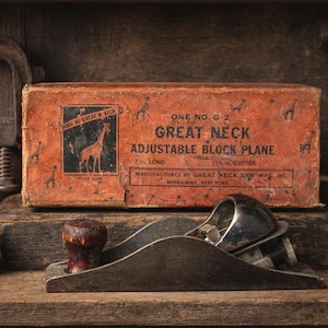 May include: A vintage adjustable block plane with a wooden handle and metal blade, on a wooden shelf. An orange box with the text "GREAT NECK ADJUSTABLE BLOCK PLANE" and a giraffe logo is behind it. Other tools are in the background.