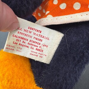 May include: A close-up of a tag attached to a stuffed toy. The tag is white with black text that reads "All New Material Contents Synthetic Fibers California Stuffed Toys 611 So. Anderson St. Los Angeles, Ca. 90023 Penn. 173 Ohio PA173 Mass. T111 Maine 56".