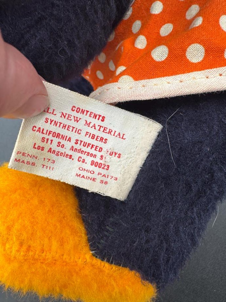 May include: A close-up of a tag attached to a stuffed toy. The tag is white with black text that reads "All New Material Contents Synthetic Fibers California Stuffed Toys 611 So. Anderson St. Los Angeles, Ca. 90023 Penn. 173 Ohio PA173 Mass. T111 Maine 56".