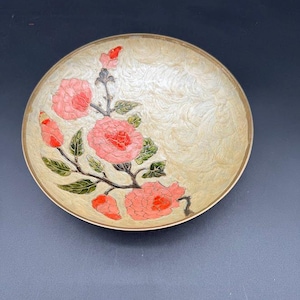 May include: A round, decorative dish with a floral design. The dish features a cream-colored background with a swirling pattern and a gold-colored rim. Pink roses and green leaves are painted on the dish.