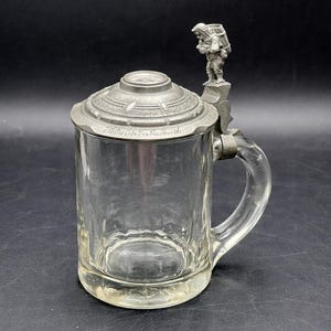 以下が含まれることがあります： 金属製の蓋と取っ手が付いた透明なガラスのビールジョッキ。 蓋にはエンボス加工のデザインと小さな置物が施されています。 取っ手は透明なガラスです。 黒い背景にあります。