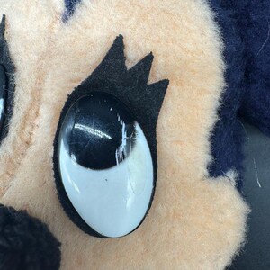 May include: Close-up of a plush toy's face. The toy has a light brown face with black eyes and eyelashes. The toy's hair is dark blue.