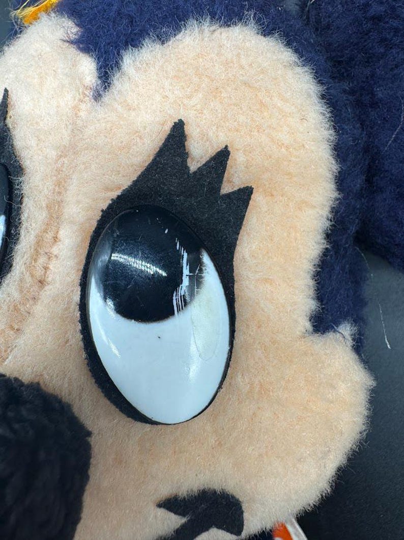 May include: Close-up of a plush toy's face. The toy has a light brown face with black eyes and eyelashes. The toy's hair is dark blue.