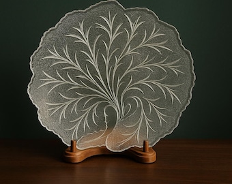 Puede incluir: Un plato decorativo de vidrio transparente con un diseño de árbol, apoyado en un soporte de madera. El plato presenta un patrón grabado de ramas y hojas. El plato tiene una forma orgánica única y se exhibe sobre una superficie de madera oscura.