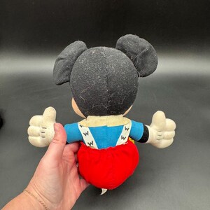 May include: A vintage Mickey Mouse plush toy, viewed from the back. The plush features black ears, a black head, a blue shirt, red shorts, and white gloves. The toy's arms are outstretched, with thumbs up.