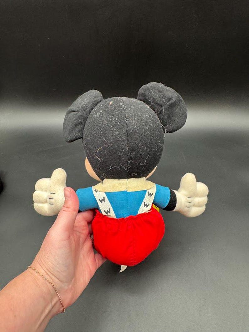 May include: A vintage Mickey Mouse plush toy, viewed from the back. The plush features black ears, a black head, a blue shirt, red shorts, and white gloves. The toy's arms are outstretched, with thumbs up.
