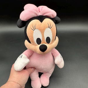May include: A plush Minnie Mouse doll in pink and black. The doll has a pink bow, a tan face with embroidered eyes and eyelashes, and black ears. The doll is wearing a pink outfit with white gloves and pink shoes.
