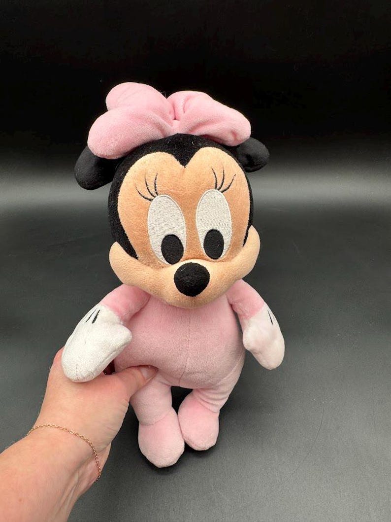 May include: A plush Minnie Mouse doll in pink and black. The doll has a pink bow, a tan face with embroidered eyes and eyelashes, and black ears. The doll is wearing a pink outfit with white gloves and pink shoes.