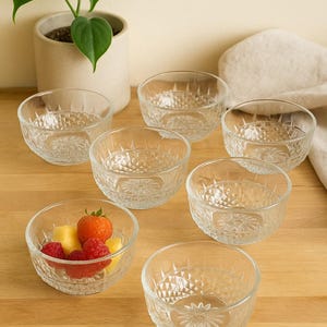 May include: A set of seven clear glass dessert bowls with a textured, starburst pattern. One bowl contains fresh fruit. A potted plant is in the background. The bowls are arranged on a wooden surface.