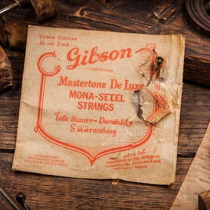 Könnte beinhalten: Vintage Gibson Mastertone De Luxe Gitarrensaiten-Packung mit rotem und cremefarbenem Text auf gealtertem Papier. Die Packung ist zerrissen und gibt die Saiten frei. Auf einer rustikalen Holzoberfläche, mit anderen Vintage-Werkzeugen.
