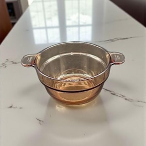 Puede incluir: Un cuenco de vidrio transparente de color ámbar con dos asas pequeñas. El cuenco tiene forma redondeada y un borde ligeramente ensanchado. Se encuentra sobre una encimera blanca, lo que sugiere su uso en la cocina o el comedor.