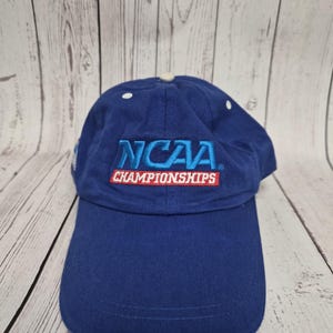 May include: A blue baseball cap with the text "NCAA CHAMPIONSHIPS" in white and red. The cap has a curved brim and a white button on top. The hat is made of a durable fabric.