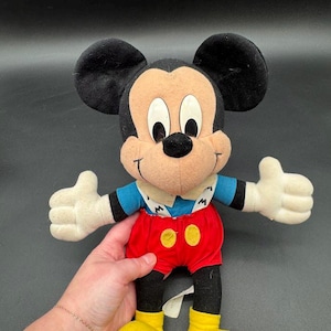 May include: A Mickey Mouse plush toy with black ears, a tan face, and white gloves. Mickey wears a blue shirt, red shorts with yellow buttons, and yellow shoes. The toy is a classic Disney character.