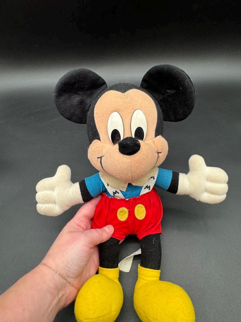 May include: A Mickey Mouse plush toy with black ears, a tan face, and white gloves. Mickey wears a blue shirt, red shorts with yellow buttons, and yellow shoes. The toy is a classic Disney character.