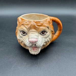 May include: A ceramic mug shaped like a tiger's head. The mug features orange and white fur details, brown eyes, and a pink nose. The handle is orange. The mug is designed to resemble a tiger's face.