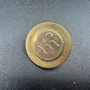 May include: A gold-toned pin with a circular design. The outer ring has a ridged pattern, while the center features a smaller circle with an ornate letter "R". The pin is set against a dark background.