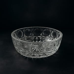Puede incluir: Un cuenco de vidrio transparente con un patrón geométrico detallado. El cuenco presenta un borde circular con diseños de círculos repetidos y un patrón central de estrella. El cuenco es de vidrio transparente y está sobre un fondo negro.