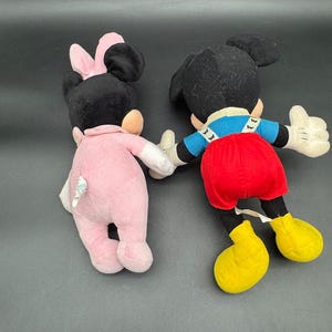 May include: Two plush Disney character dolls, Mickey Mouse and Minnie Mouse, are shown from the back. Mickey wears red shorts, yellow shoes, and a blue shirt. Minnie is in a pink outfit with a pink bow.