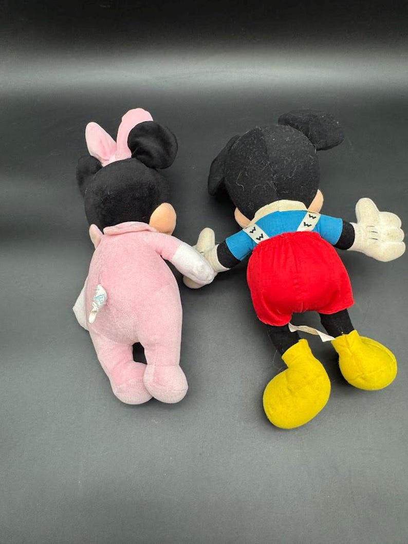 May include: Two plush Disney character dolls, Mickey Mouse and Minnie Mouse, are shown from the back. Mickey wears red shorts, yellow shoes, and a blue shirt. Minnie is in a pink outfit with a pink bow.