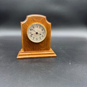 Puede incluir: Un pequeño reloj de repisa de madera con un acabado marrón claro. La esfera del reloj es redonda con un bisel dorado y números negros. El reloj tiene una base rectangular y una parte superior decorativa.