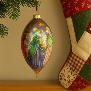 May include: An oval Christmas bauble with a religious theme. The bauble depicts three figures in colourful robes, holding gifts and a golden chalice. The bauble is suspended from a red ribbon, with a Christmas stocking in the background.