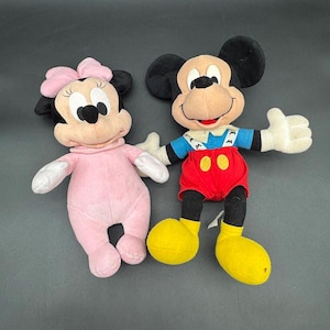 May include: Two plush Disney character dolls: Mickey Mouse and Minnie Mouse. Mickey wears red shorts, yellow shoes, and a blue shirt. Minnie is in a pink outfit with a matching bow. Both have black ears and white gloves.