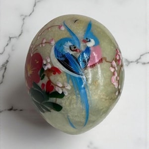 May include: A hand-painted, oval-shaped decorative stone. The stone features a design of two blue and pink birds perched on a branch with pink blossoms. Red flowers and green leaves are also painted on the stone.