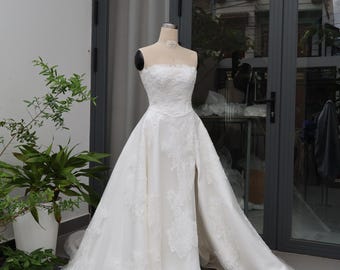 May include: A strapless white wedding dress on a mannequin. The gown features intricate lace detailing on the bodice and skirt, with a high slit on the front. The dress is displayed in front of a glass door.