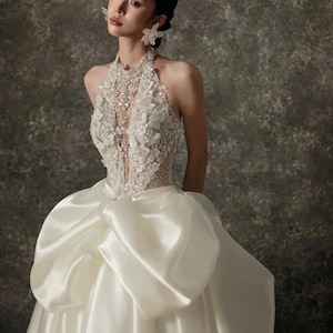 May include: A white satin wedding gown with a halter neckline and a full skirt. The bodice is embellished with floral lace and sparkling crystals. The model is wearing flower-shaped earrings.