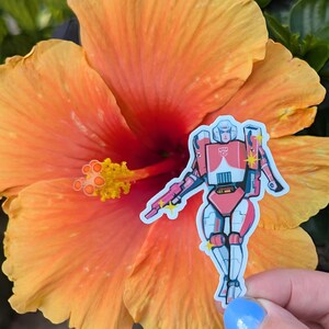 Puede incluir: Una pegatina troquelada que muestra un personaje robot estilizado en tonos rosa, blanco y azul claro. El robot tiene una insignia Autobot roja en el pecho, sostiene un bláster rosa y está decorado con pequeños detalles de estrellas amarillas.