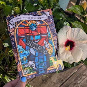 May include: A rectangular art print featuring a stained-glass style illustration of a red and blue robot holding a large black blaster. The text "BE STRONG ENOUGH TO BE GENTLE" is printed at the top. The design includes white doves and a golden deer.