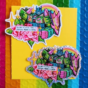 Könnte beinhalten: Zwei Aufkleber mit einem Roboterdesign in Grün, Blau und Pink, mit einem Regenbogen und rosa Blumen. Der Text auf dem Aufkleber lautet "YOU ARE A LITTLE SOUL CARRYING AROUND A CORPSE". Die Aufkleber befinden sich auf einem gelben Hintergrund.
