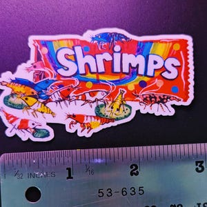 May include: A colorful sticker featuring the word "Shrimps" in white letters on a red and yellow background. The sticker showcases various shrimp illustrations in shades of red, yellow, blue, and black. A ruler is placed below the sticker.