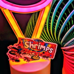 May include: A colorful toy with a rainbow slinky and a neon pink and yellow frame. A sticker with the word "Shrimps" and illustrations of shrimp is attached to the toy.
