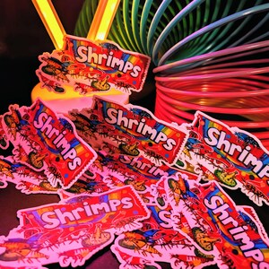 May include: A collection of colorful stickers with the word "Shrimps" in a rainbow banner. The stickers feature illustrations of shrimp and are scattered on a dark surface, with a rainbow slinky and a yellow light in the background.