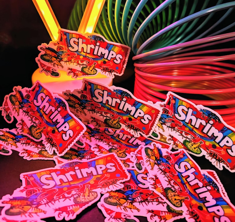 May include: A collection of colorful stickers with the word "Shrimps" in a rainbow banner. The stickers feature illustrations of shrimp and are scattered on a dark surface, with a rainbow slinky and a yellow light in the background.