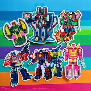 May include: A selection of robot character stickers in various colours. The stickers depict robot designs in red, blue, green, yellow, and pink. One sticker has the text "ALL HAIL STAR SCREAM". The stickers are on a rainbow striped background.