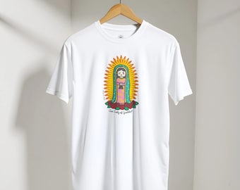 May include: A white short-sleeve t-shirt featuring a central graphic of a stylized, cartoon-like Our Lady of Guadalupe figure surrounded by a yellow sunburst, standing on red roses, with the text Our Lady of Guadalupe printed below.