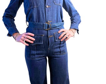 Puede incluir: Un mono de mezclilla azul oscuro con camisa de cuello, mangas largas y cinturón. El mono tiene varios bolsillos y cierre de botón.