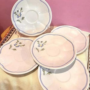 Pu&ograve; includere: Set di quattro piattini in ceramica bianca con un motivo floreale blu e verde. I piattini hanno un bordo ondulato e una fascia blu intorno al bordo.