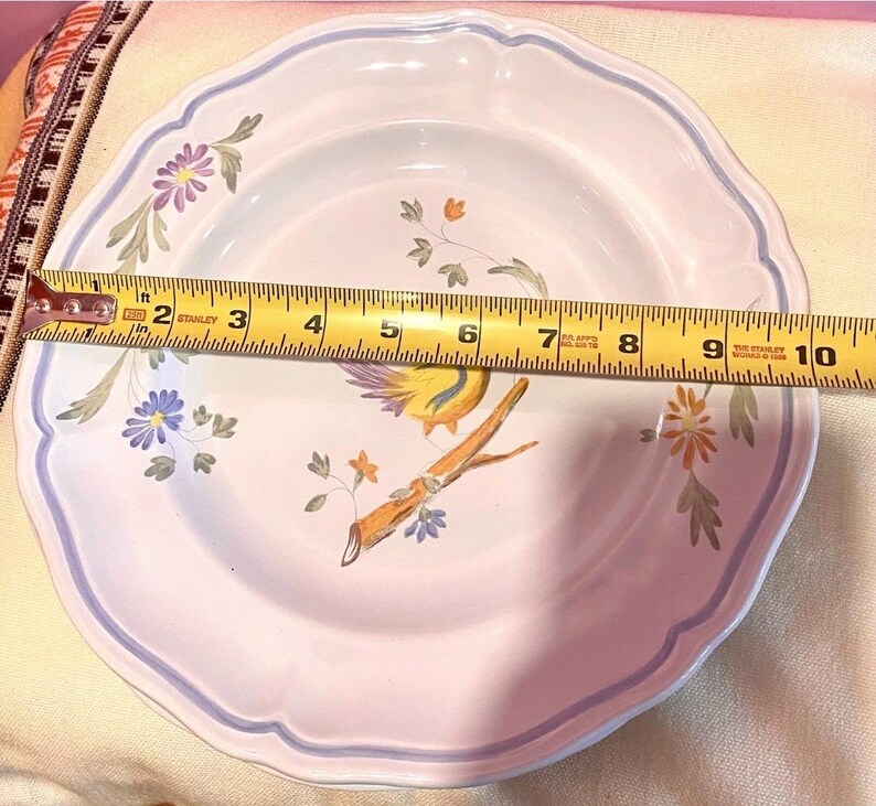 May include: A white ceramic plate with a scalloped edge and a blue border. The plate features a painted design of a yellow bird perched on a branch with flowers. The plate measures approximately 10 inches in diameter.